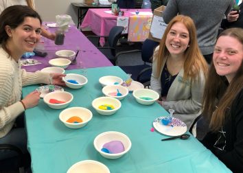 Three smiling students sit a table with bowls of paint.