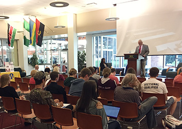 A man in a suit stands at a podium in front of an audience in the Student Union at SUNY Jamestown Community College. There are four international flags hanging on the wall to his right.