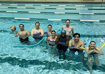 Students holding pool noodles and a soccer ball while standing in a pool with lane lines floating in the pool behind them.