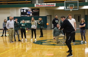 Students playing games in gymnasium. One is a batter, the rest are behind him.
