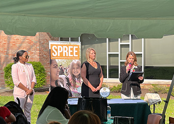 Three women stand before an audience to present awards at a picnic.