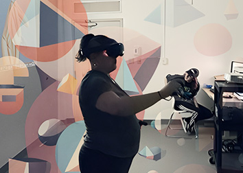 A student stands in a room using a Virtual Reality headset and controllers while another student sits at a table with a computer in the corner, and looks back at a screen projected on the wall. The image has been enhanced by artificial intelligence to include geometric shapes in the air around the students.