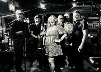 A group of six people holding different types of horns in a music studio with spotlights above them.