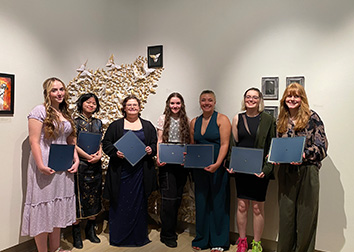 Art students stand in the Weeks Gallery holding certificates with works of art on the walls behind them.