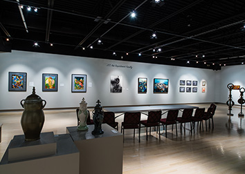 Lighted works of art line a gallery wall in the background with a table and row of chairs in the mid-ground, and a sculpture in the shadows on the left side of the foreground.