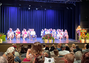 People sitting in chairs on a stage looking at a person standing at a podium with plants decorating the front of the stage and a crowd filling the seats in the theater.