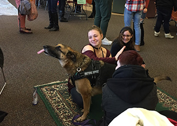 Students sit on a floor while petting a dog with a harness that says "Emotional Support."