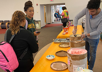 Students stand in front of a table with tubs of ice cream and another student scooping.