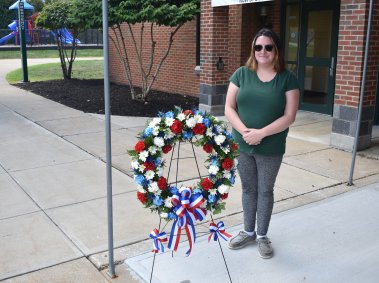 A woman stands next to a patriotic floral wreath with red, white, and blue flowers and ribbons, placed on a stand outside a brick building.