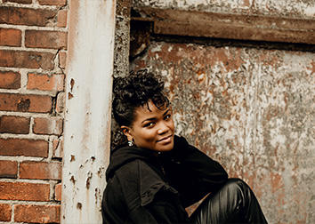 A woman leans against a doorway framed by a brick wall.