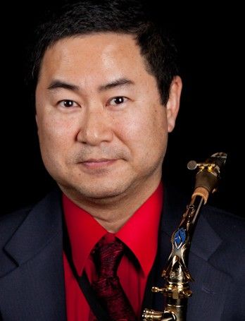 A man in a dark suit and red shirt holds a saxophone against a black background.