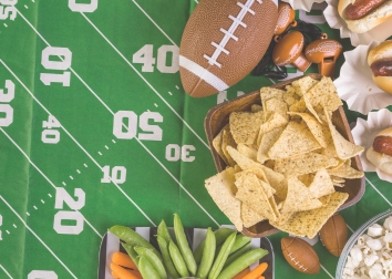 A table decorated with a miniature football field table cover and a football, chips and veggies set along the right side.