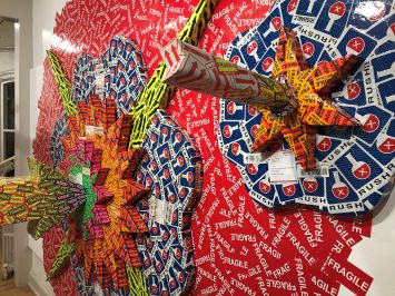 Colorful 3D wall art made of layered shipping labels, including "Fragile" and "Rush." The artwork features symmetrical starburst patterns creating a vibrant and dynamic effect.