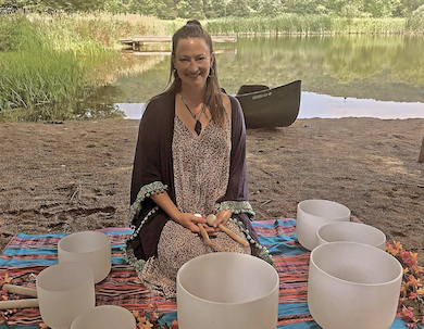 A person kneeling on a blanket in a sandy area with a body of water and canoe in the background.