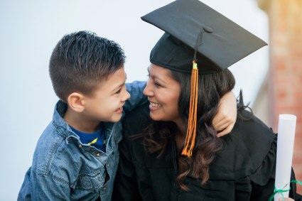 A mother wearing graduation cap and gown hugs her child while smiling.