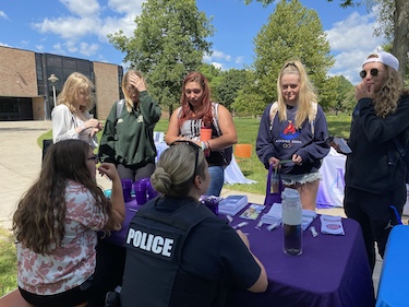 Students stand in front of a table talking to police representatives outside on a college campus.