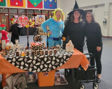 Three people in Halloween costumes stand behind a table decorated for a Halloween event, with crafts, candy, and festive banners in the background.