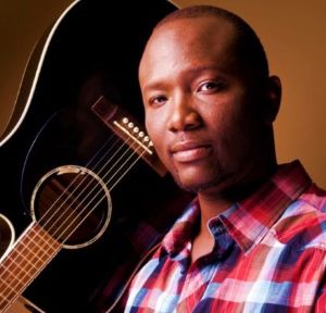 Man holding a guitar up side down on his right shoulder while looking directly at the camera.
