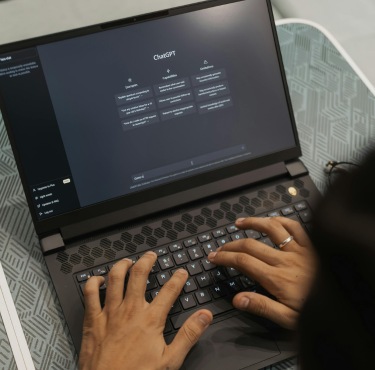 Hands typing on a laptop with ChatGPT on the screen. The setting is informal, with a patterned surface in the background.