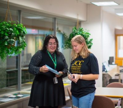 Two women in an office setting look at documents together. One wears a black dress, the other a T-shirt and jeans. Green plants hang above them.