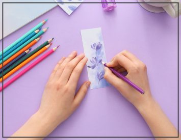 A person draws purple flowers on a paper bookmark. with a colored pencil. Colorful pencils nearby on a lavender surface.