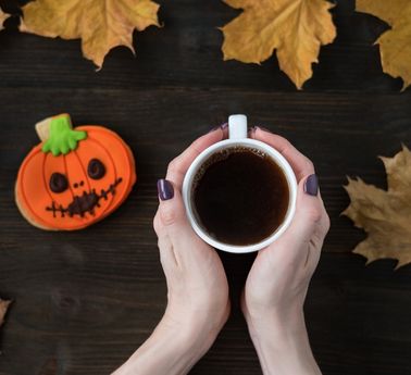Hands holding a white mug of coffee, with a pumpkin-shaped Halloween cookie and autumn leaves on a dark wooden background.
