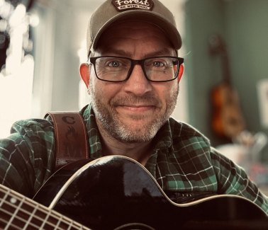 A man wearing a baseball cap and plaid shirt holds a guitar, looking directly at the camera.