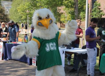 A person in an JCC Jayhawk mascot costume at an outdoor event. The mascot is wearing a green jersey with "JCC" on it.