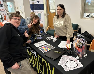 Three people stand by a table with "National Comedy" materials. One person gives a thumbs-up, another makes a peace sign, and a woman smiles warmly.