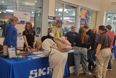 A group of people stand around a table with various items displayed at an event. A woman in a blue shirt and a man in a safety vest are in the foreground.