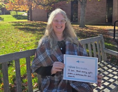 A smiling woman sits on a wooden bench in a sunny autumn courtyard, holding a handwritten “I Give Because” sign; the scene feels warm and hopeful.
