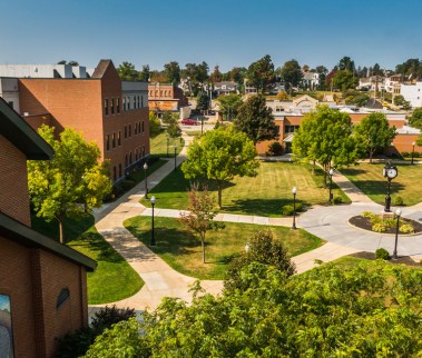 Aerial view of a college campus quad with brick buildings, pathways, and lush green trees. The scene is bright and peaceful, conveying a serene academic atmosphere.
