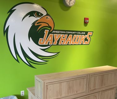Large stylized eagle logo and "Jamestown Community College Jayhawks" text on a bright green wall above a wooden reception desk.