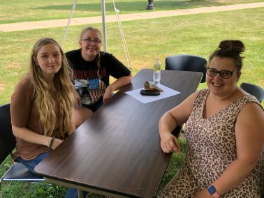 Three smiling individuals sit at a wooden table under a white tent, with a grassy outdoor area in the background.