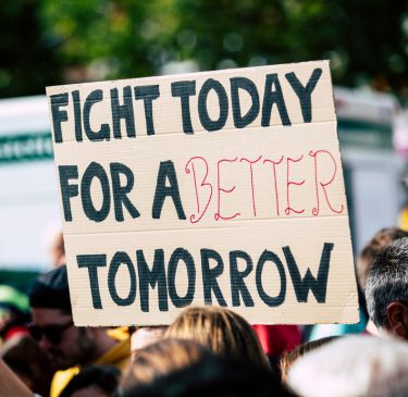 A hand holds a protest sign reading "Fight Today for a Better Tomorrow" in bold letters. The scene is vibrant, suggesting hope and activism in a crowded rally.