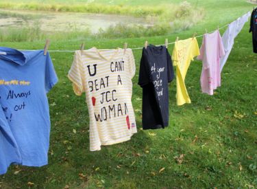 Laundry hangs on a clothesline in front of a grassy field and a body of water. T-shirts of various colors and designs are displayed.