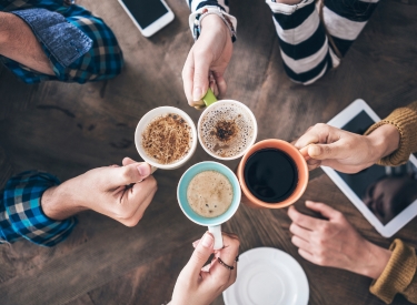 Overhead view of a group of people's hands reaching for mugs of coffee and a tablet. The image captures a casual gathering with warm drinks and technology.