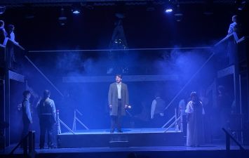 A lone actor stands center stage under blue lighting, surrounded by fog. Several actors in the background create a dramatic, mysterious scene.
