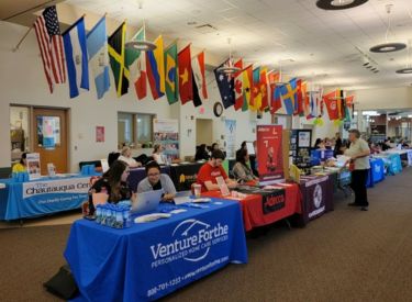 A college career fair with rows of tables displaying flags and informational materials. People are gathered around tables, interacting with representatives and students.