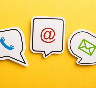 Three speech bubbles on a bright yellow background show a blue phone handset, red @ symbol, and green envelope, representing digital communication.