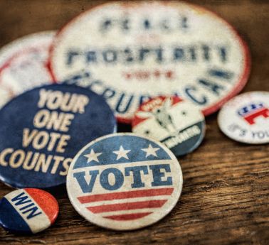 Vintage political buttons on a wooden surface, featuring “VOTE,” “Your One Vote Counts,” and patriotic red, white, and blue designs.