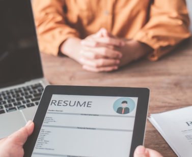 Tablet displaying a resume with a person's hands holding it, in front of a person sitting at a wooden table with a laptop and papers.