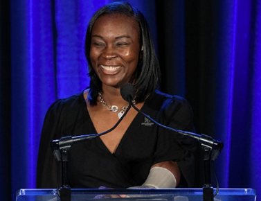 A cheerful woman in a black dress speaks at a podium against a backdrop of blue curtains. Her confident smile conveys a positive, engaging tone.