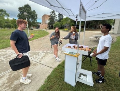 Four people are gathered around an outdoor tent stand displaying donuts on a campus walkway.