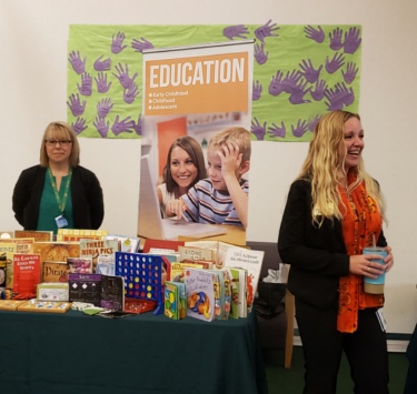 Two women stand near a table filled with children's books. A banner with "Education" notes early childhood to adolescent focus.
