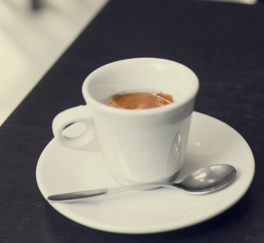 A small white espresso cup with coffee sits on a matching saucer and spoon on a dark table, creating a calm, understated café scene.