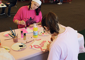 Students sit across from each other at a table. They are painting on small art boards and there are paper plates with paints and cup full of brushes and paint-stained water on the table.
