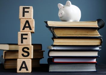 Wood blocks with letters stacked to form the word FAFSA next to stacks of books with a white piggy bank on top one of the book stacks.