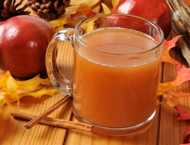 A clear glass mug filled with amber-colored apple cider, surrounded by red apples, cinnamon sticks, pinecones, and colorful autumn leaves on a wooden surface.