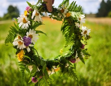 A hand holds a vibrant floral wreath against a lush, green meadow. The wreath features daisies, purple flowers, and fern leaves, evoking a summery, cheerful mood.